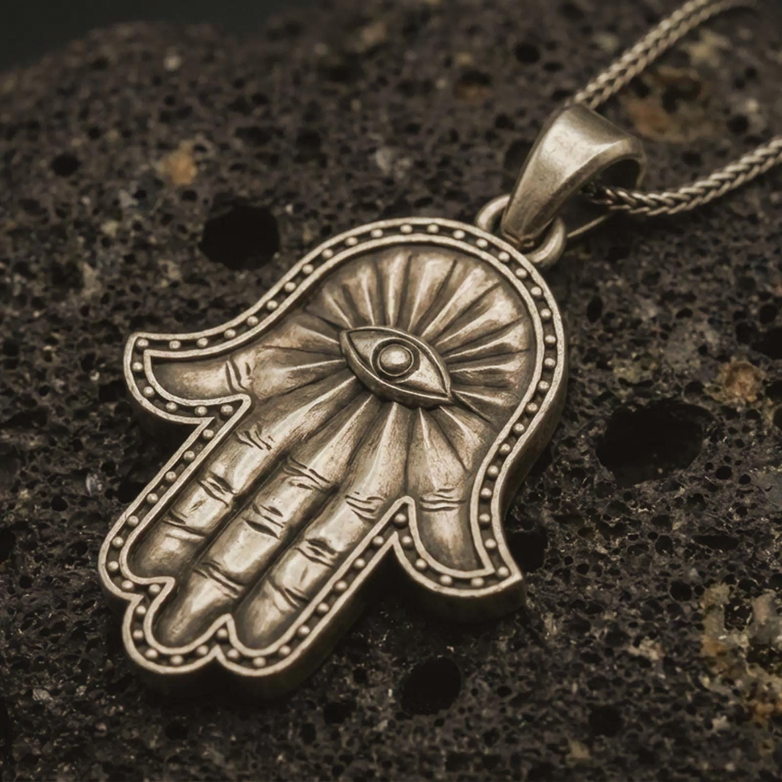 Bronze Hamsa hand pendant necklace with an oxidized finish resting on textured volcanic rock, captured in a close-up premium studio composition.
