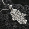 Load image into Gallery viewer, Polished silver Jerusalem Hamsa hand pendant necklace displayed on black volcanic rock, shown in a refined macro product shot with Hebrew inscription and detailed cityscape relief.
