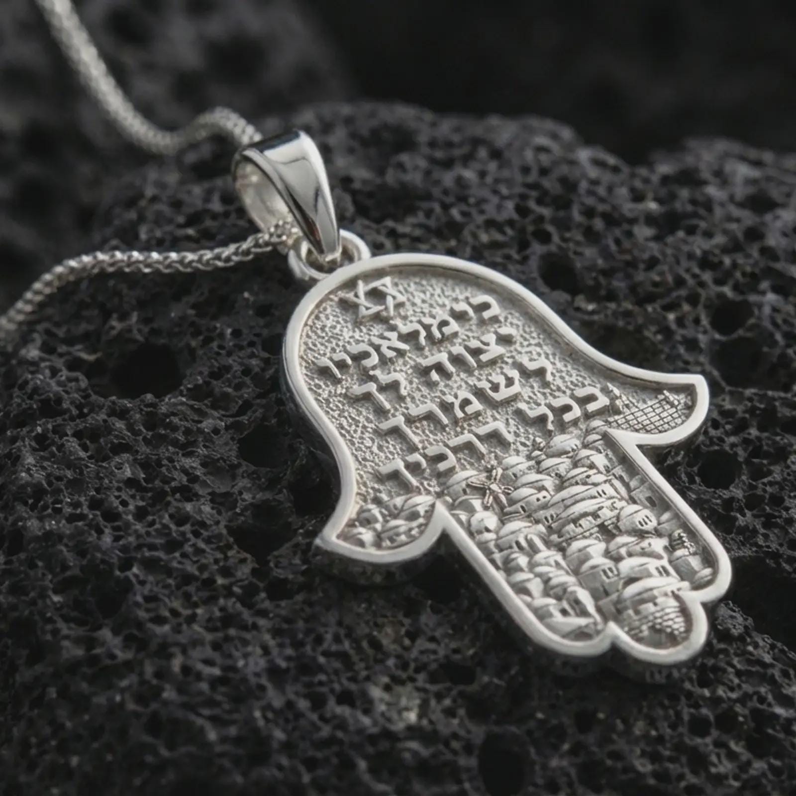 Polished silver Jerusalem Hamsa hand pendant necklace displayed on black volcanic rock, shown in a refined macro product shot with Hebrew inscription and detailed cityscape relief.
