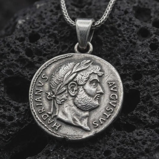 Oxidized Emperor Augustus coin pendant necklace resting on black volcanic stone, featuring a Roman imperial side profile with laurel wreath and engraved ancient lettering in a detailed macro product photograph.