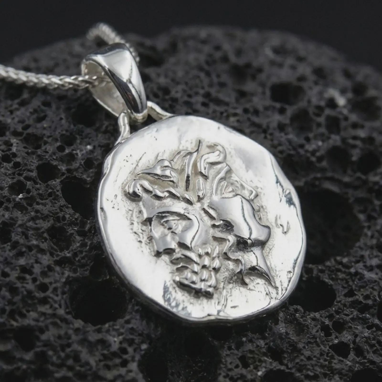 Polished silver Janus coin pendant necklace displayed on dark volcanic rock, shown in a premium macro product shot that highlights the dual face motif and worn classical medallion texture.