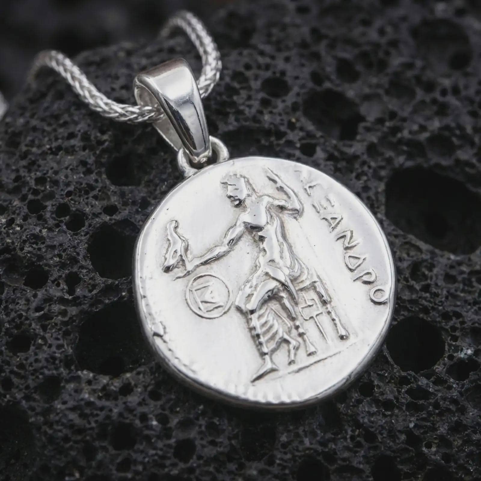 Polished silver Alexander the Great coin pendant necklace displayed on dark volcanic rock, shown in a premium close-up image that highlights the antique medallion texture, heroic figure, and historical Greek inscription.