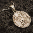 Load image into Gallery viewer, Bronze Alexander the Great coin pendant necklace resting on black volcanic stone, featuring an ancient Greek coin-style relief with a standing warrior figure and engraved classical lettering in a detailed macro product photograph.
