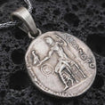 Load image into Gallery viewer, Oxidized Alexander the Great coin pendant necklace resting on black volcanic stone, featuring an ancient Greek coin-style relief with a standing warrior figure and engraved classical lettering in a detailed macro product photograph.
