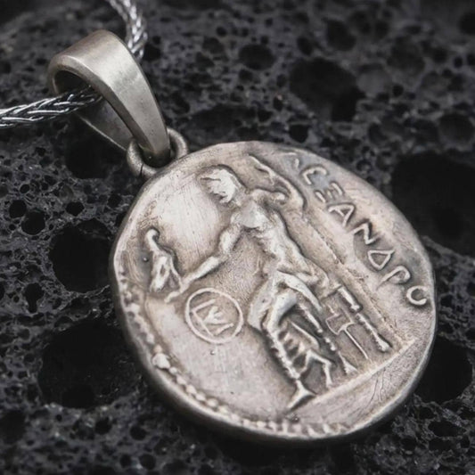 Oxidized Alexander the Great coin pendant necklace resting on black volcanic stone, featuring an ancient Greek coin-style relief with a standing warrior figure and engraved classical lettering in a detailed macro product photograph.