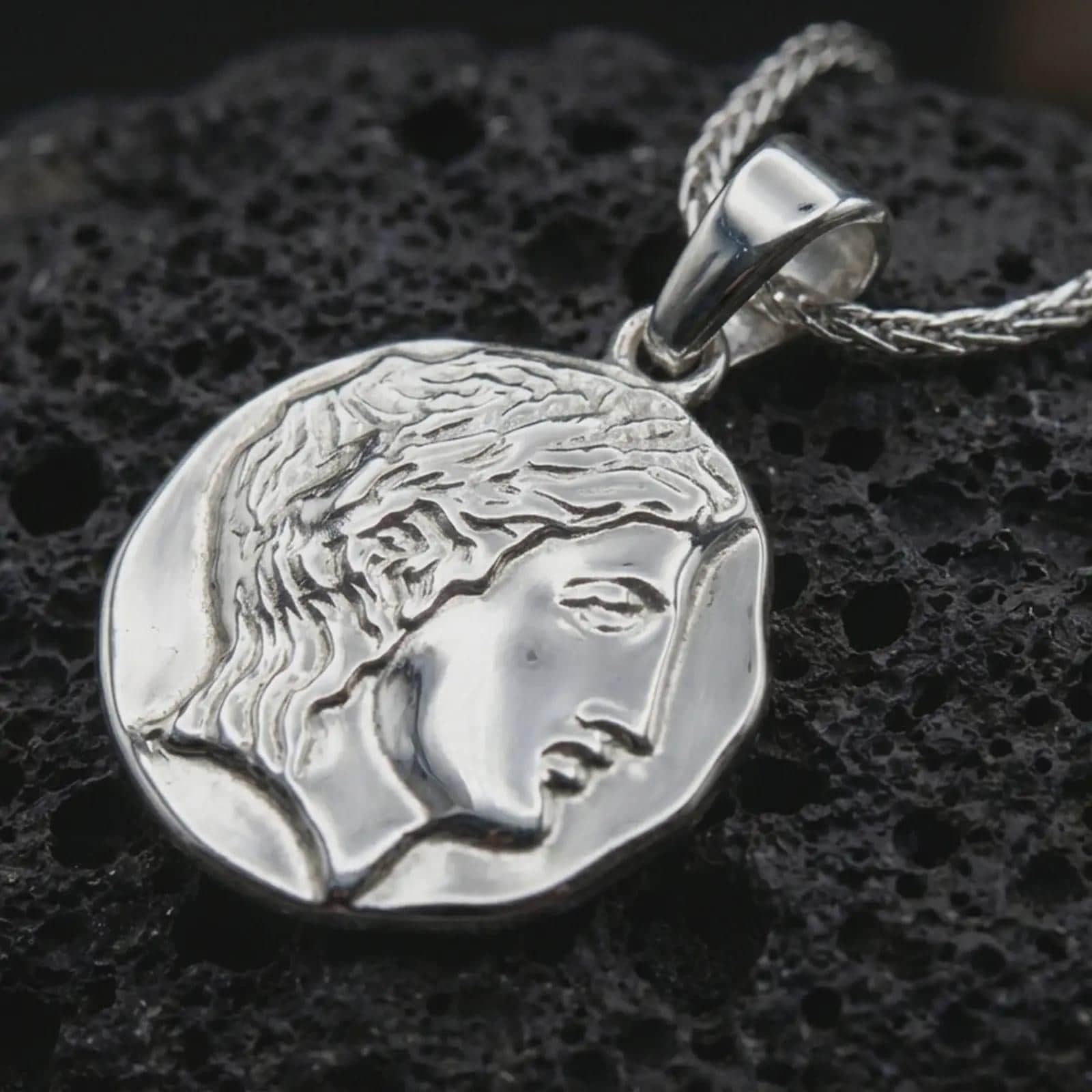Polished silver ancient Greek coin pendant necklace displayed on dark volcanic rock, shown in a premium macro product shot that highlights the feminine side profile and sculpted coin edge.