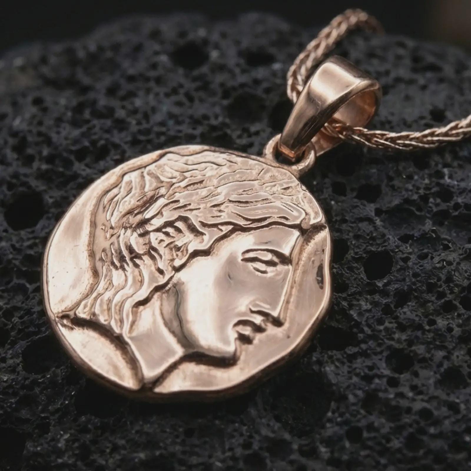 Rose gold plated ancient Greek coin pendant necklace on textured volcanic rock, showcased in a luxury macro composition with the elegant female face and weathered ancient medallion form.