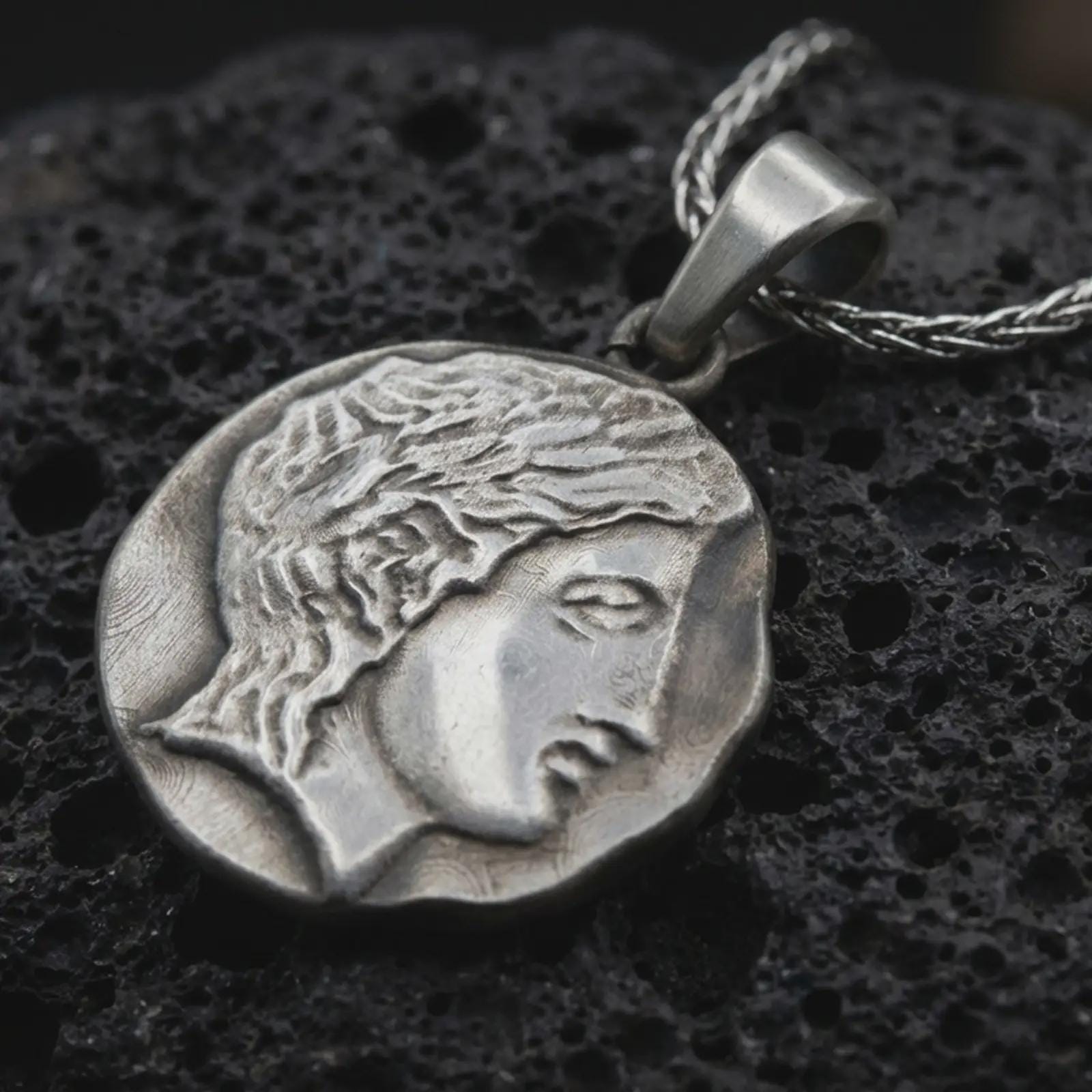 Oxidized ancient Greek coin pendant necklace resting on black volcanic stone, featuring a classical female profile in an irregular coin-shaped relief with antique-inspired surface texture.