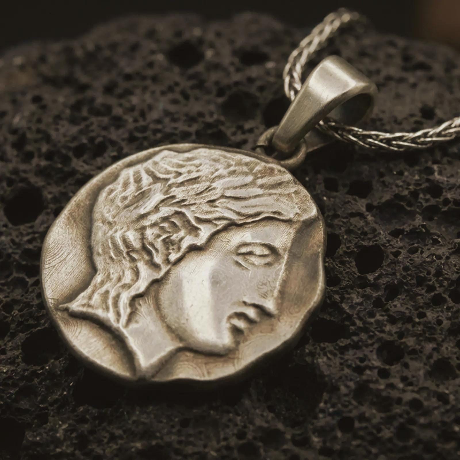 Bronze ancient Greek coin pendant necklace resting on black volcanic stone, featuring a classical female profile in an irregular coin-shaped relief with antique-inspired surface texture.