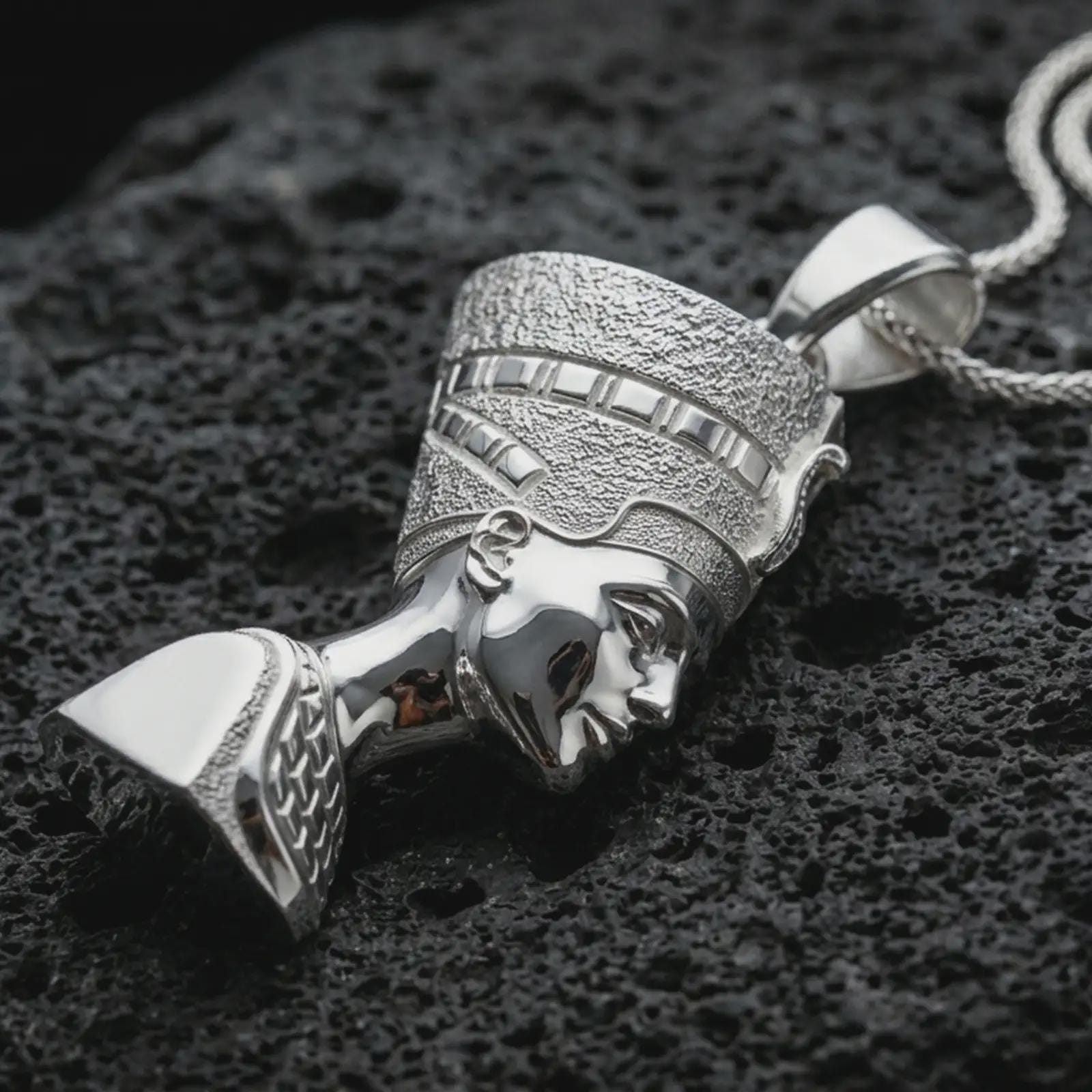 Polished silver Nefertiti pendant necklace displayed on black volcanic rock, shown in a refined macro product shot with regal Egyptian queen profile, detailed crown, and sculptural bust relief.