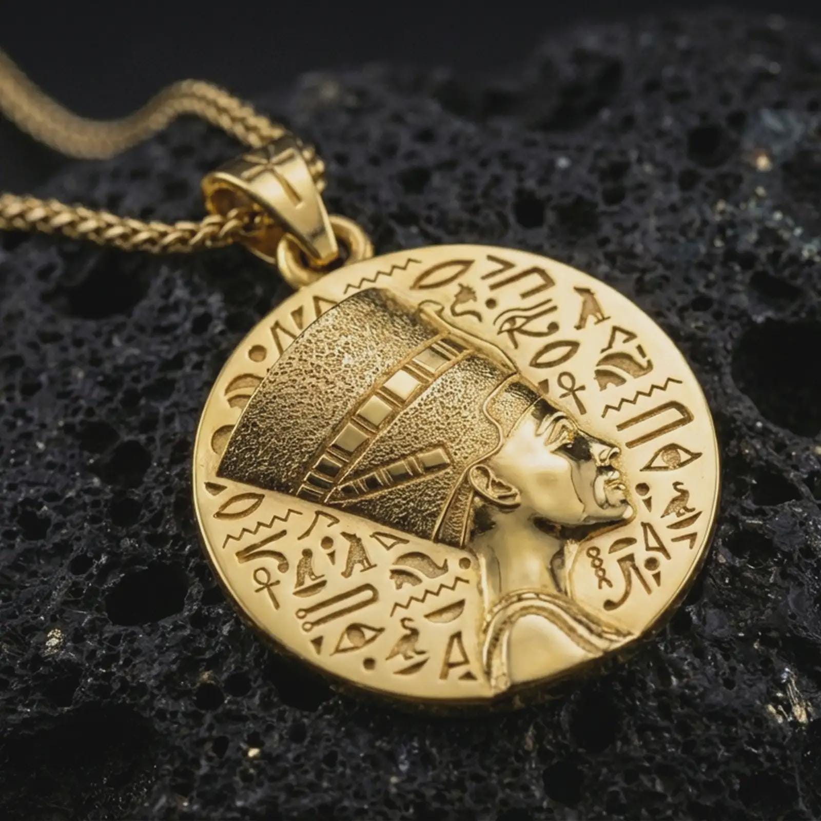 Gold plated Nefertiti medallion pendant necklace presented on porous volcanic rock, highlighting the ancient Egyptian portrait and circular coin-style design in a luxurious close-up jewelry image.