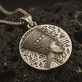 Load image into Gallery viewer, Bronze Nefertiti medallion pendant necklace with an oxidized finish resting on textured volcanic rock, captured in a close-up premium studio composition.
