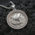 Load image into Gallery viewer, Polished silver Viking ship pendant necklace displayed on black volcanic rock, shown in a refined macro product shot with longship sail, ocean waves, and Celtic border detail.
