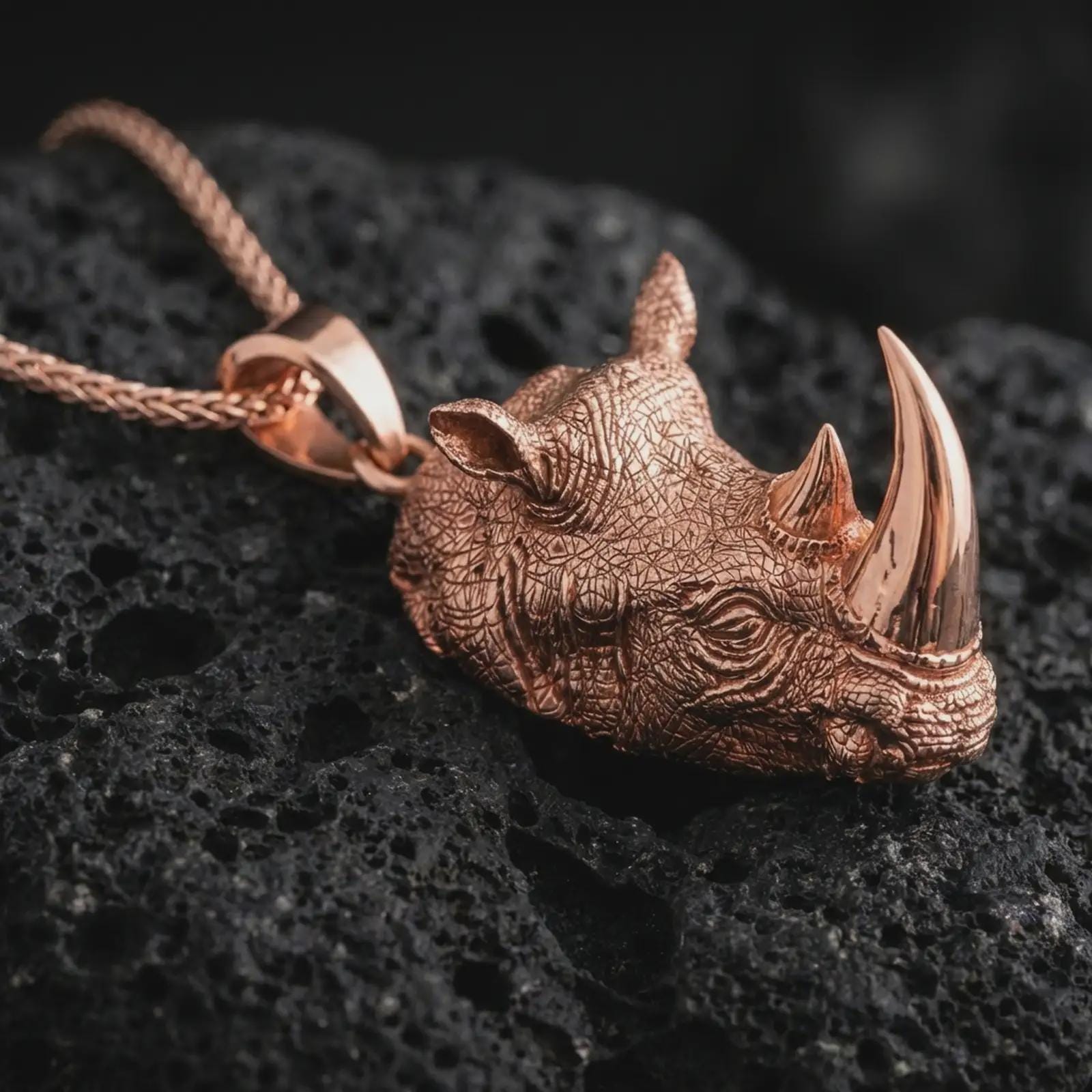 Rose gold plated rhinoceros head pendant necklace on dark volcanic rock, photographed in a detailed premium studio macro view.