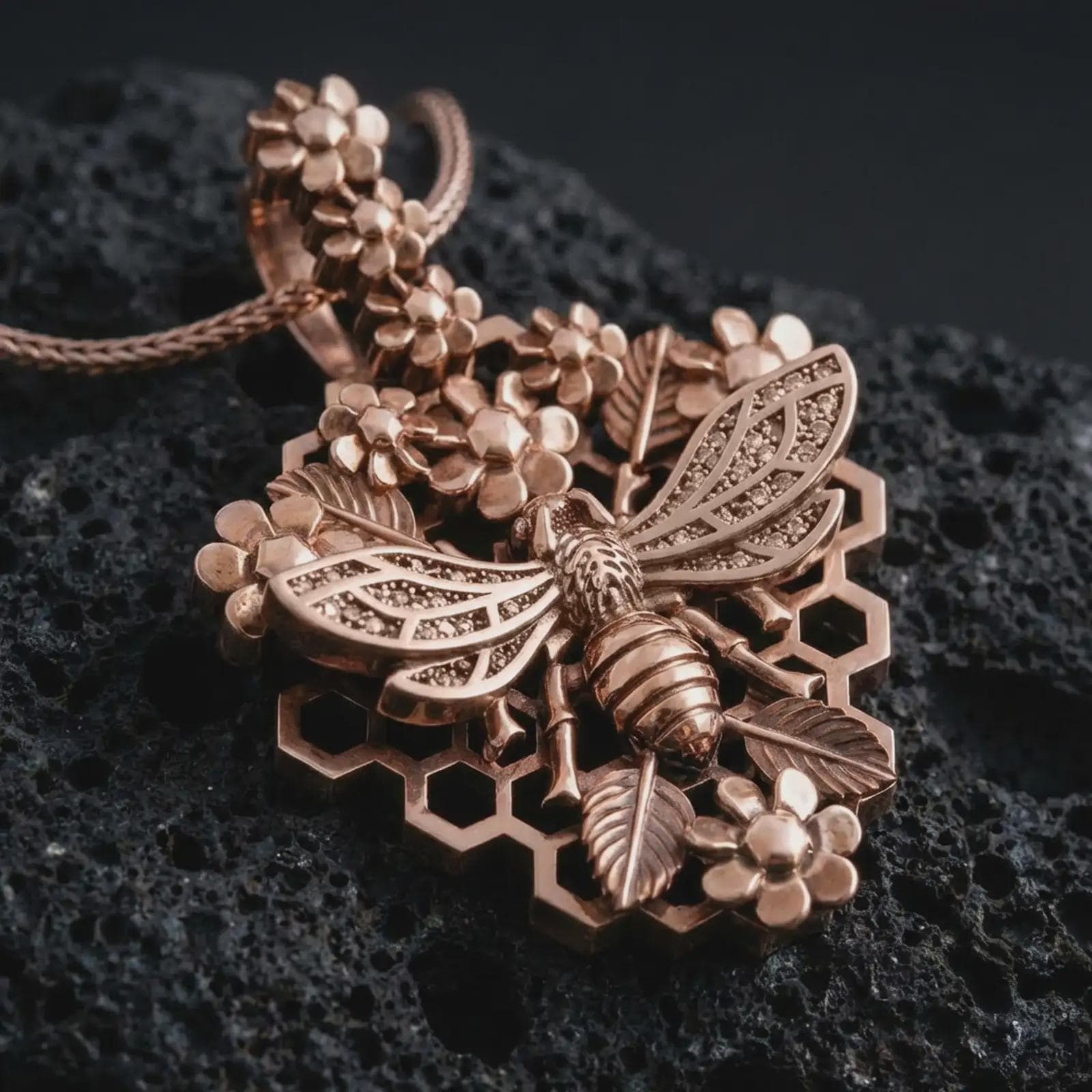 Rose gold plated floral bee pendant necklace on dark volcanic rock, photographed in a detailed premium studio macro view with botanical accents and geometric honeycomb details.