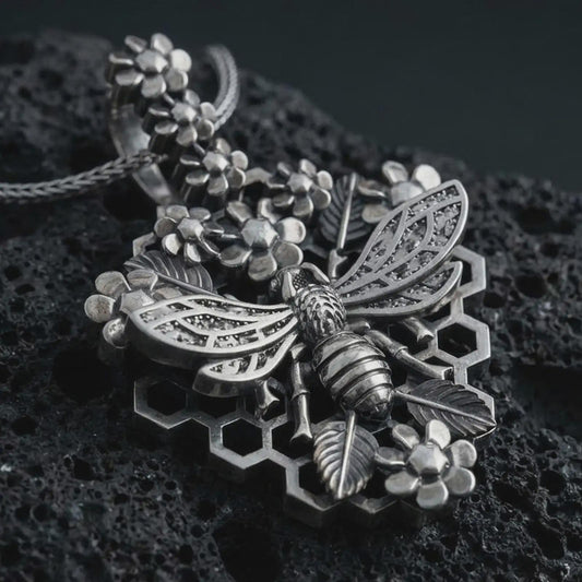 Sterling silver floral bee pendant necklace with an oxidized finish resting on textured volcanic rock, captured in a close-up premium studio composition with honeycomb, blossoms, and detailed wing relief.