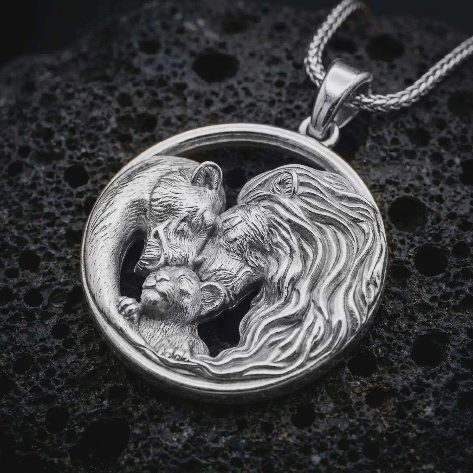 Polished silver lion and bear family pendant necklace displayed on black volcanic rock, shown in a refined macro product shot with intertwined adult animals, cub detail, and circular unity frame.