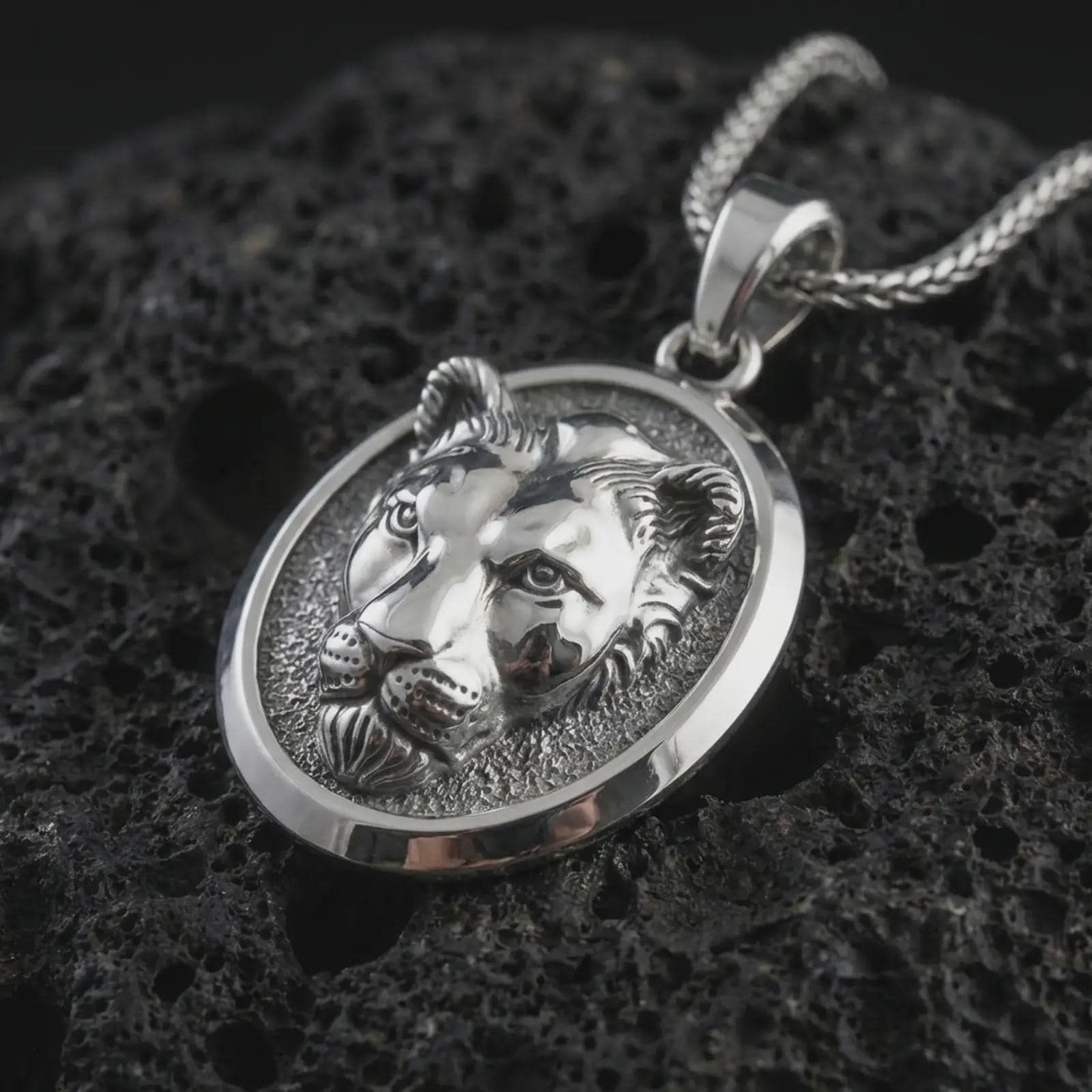 Polished silver lioness pendant necklace displayed on black volcanic rock, shown in a refined macro product shot with feline face relief, smooth oval medallion frame, and powerful wildlife detail.