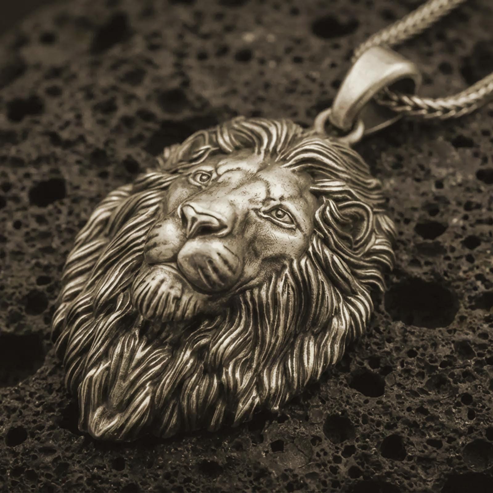 Bronze lion head pendant necklace with an oxidized finish resting on textured volcanic rock, captured in a close-up premium studio composition.