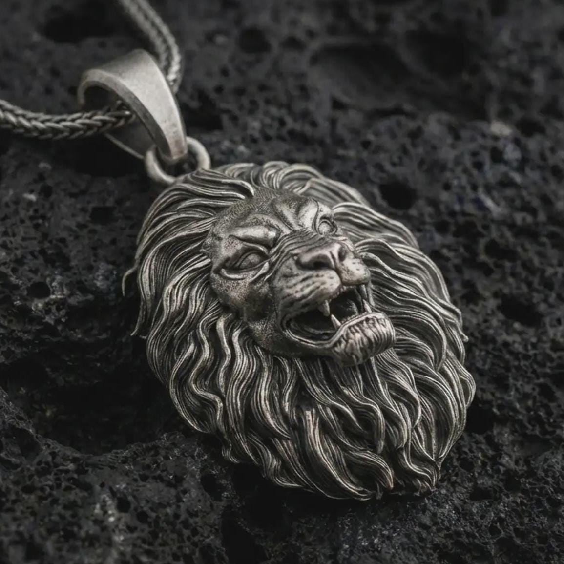 Sterling silver roaring lion head pendant necklace with an oxidized finish resting on textured volcanic rock, captured in a close-up premium studio composition.