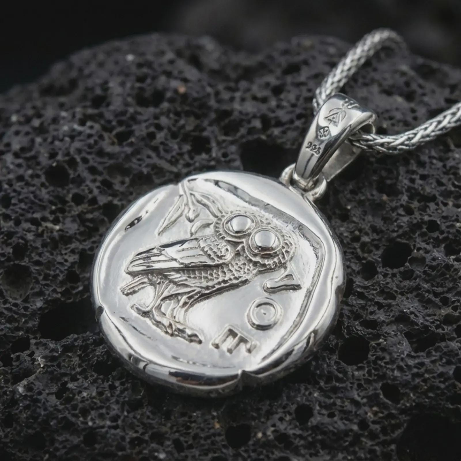 Polished silver Athena owl coin pendant necklace displayed on dark volcanic rock, shown in a premium close-up image that highlights the sacred owl motif and antique coin texture.