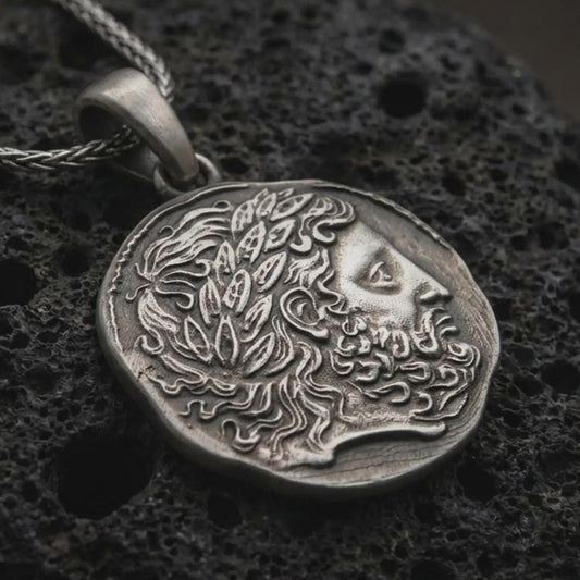 Oxidized ancient Greek coin pendant necklace resting on black volcanic stone, featuring a bearded classical profile with flowing hair and laurel-like ornament in a detailed macro product photograph.