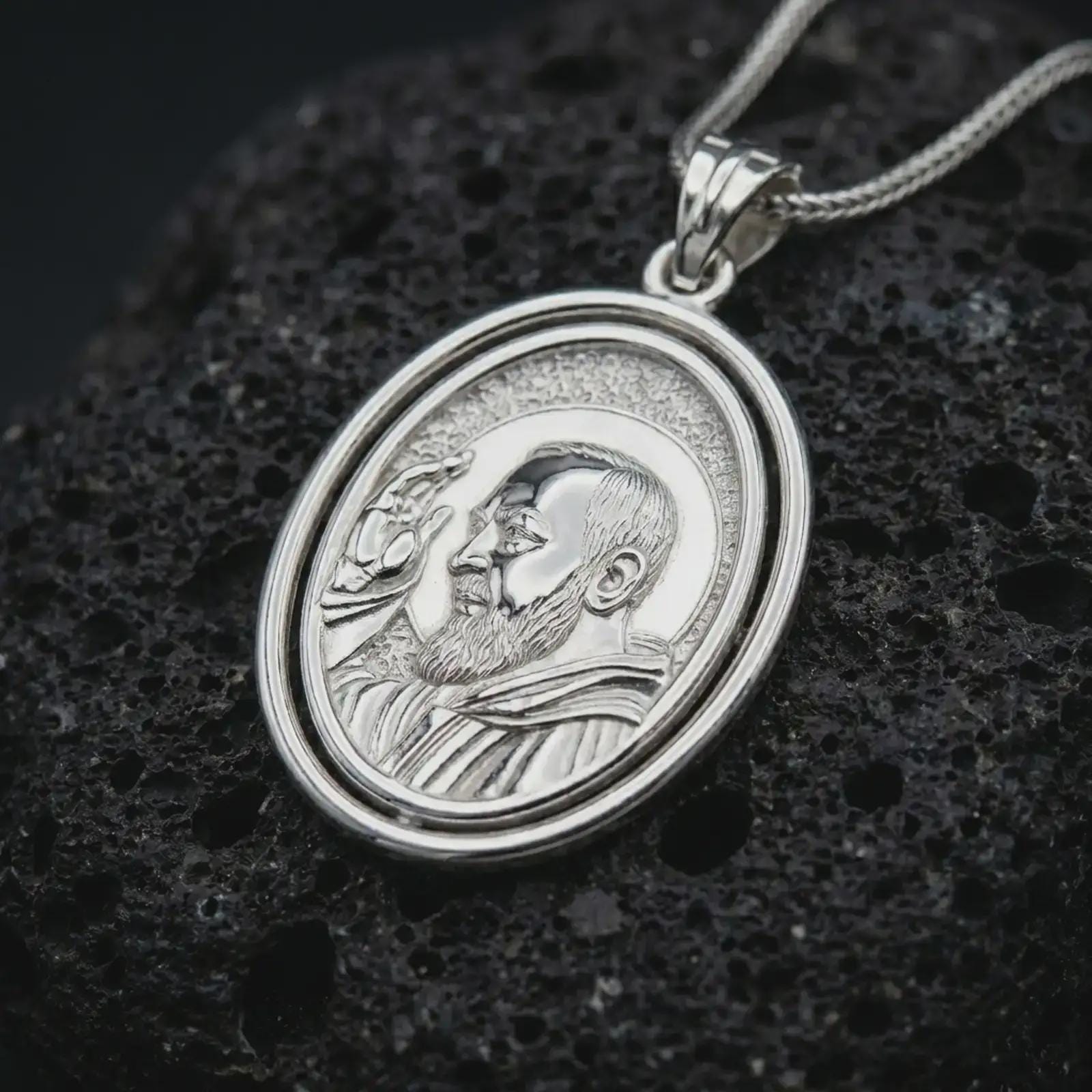 Polished silver Saint Padre Pio spinning pendant necklace displayed on dark volcanic rock, shown in a premium close-up image that highlights the saint portrait, halo, and layered oval frame.