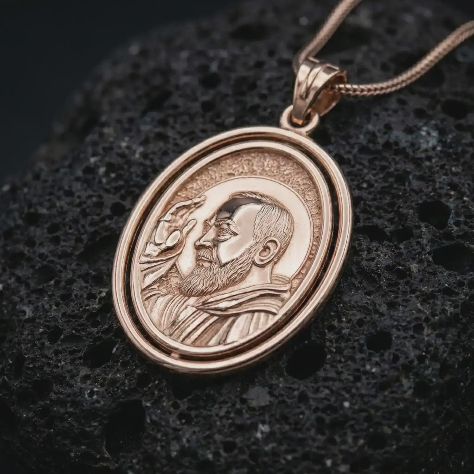 Rose gold plated Saint Padre Pio spinning pendant necklace presented on textured volcanic stone, showcasing the sacred portrait, oval spinner structure, and refined devotional detailing in a macro studio shot.