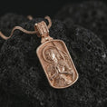 Load image into Gallery viewer, Rose gold plated Joan of Arc dog tag pendant necklace presented on textured volcanic stone, showcasing the haloed saint portrait, prayerful pose, and refined devotional detailing in a macro studio image.
