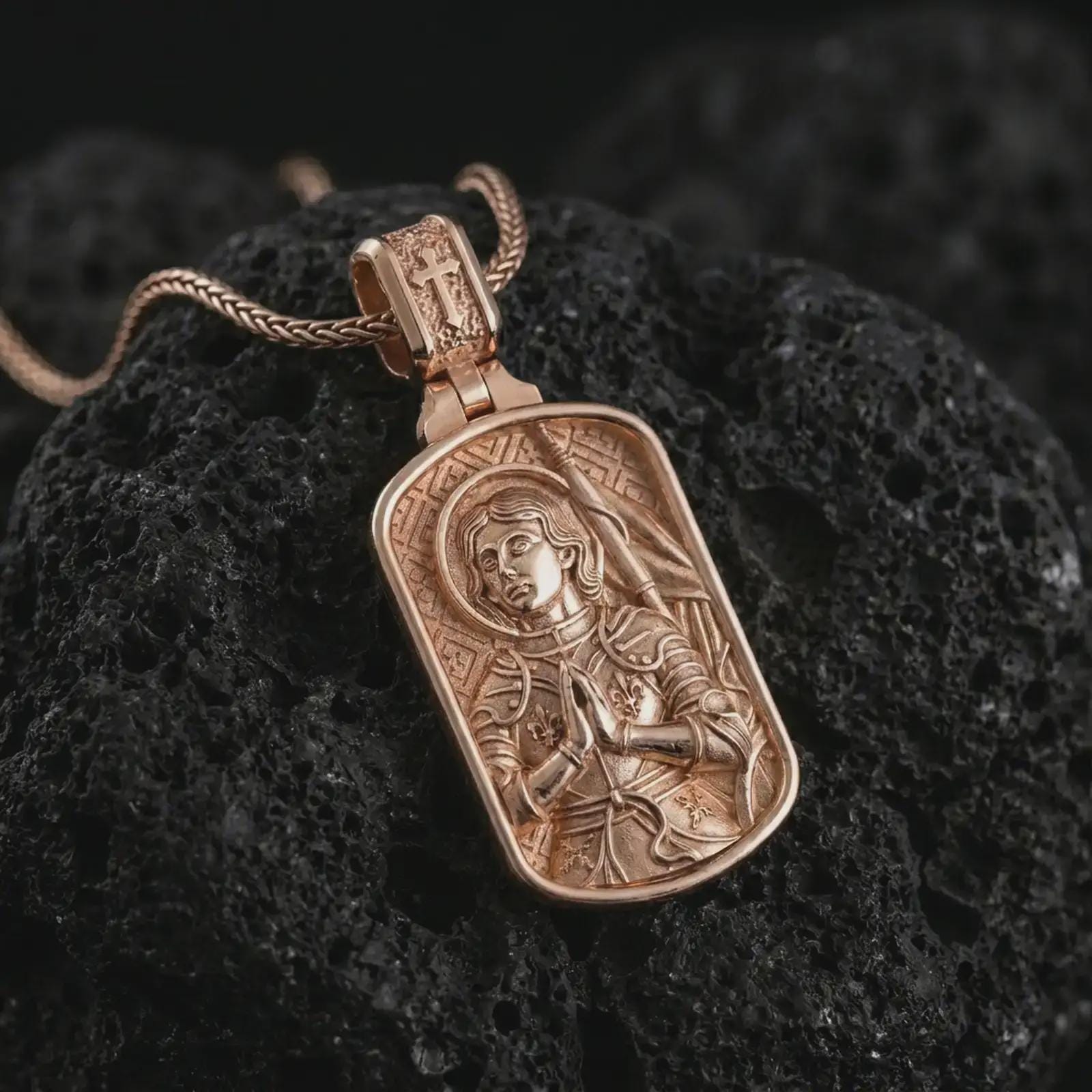 Rose gold plated Joan of Arc dog tag pendant necklace presented on textured volcanic stone, showcasing the haloed saint portrait, prayerful pose, and refined devotional detailing in a macro studio image.