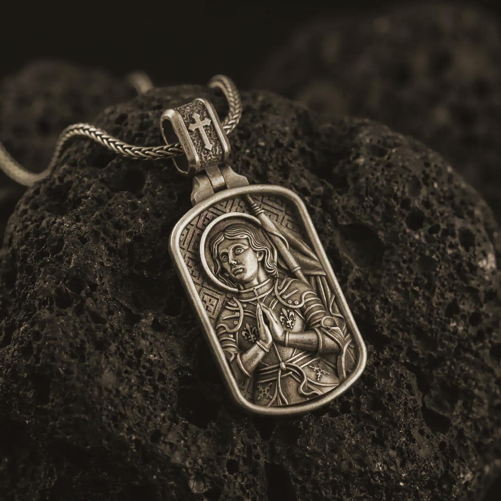 Bronze Joan of Arc dog tag pendant necklace resting on black volcanic stone, featuring a detailed saint warrior portrait with halo, folded hands, and armor in a devotional sterling silver design.