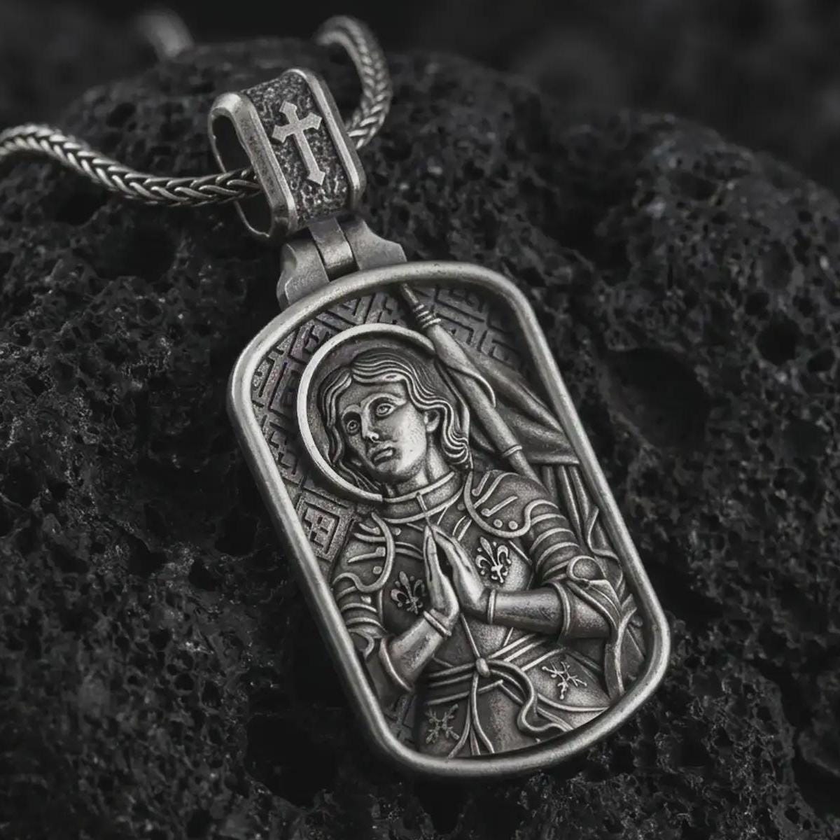 Oxidized Joan of Arc dog tag pendant necklace resting on black volcanic stone, featuring a detailed saint warrior portrait with halo, folded hands, and armor in a devotional sterling silver design.
