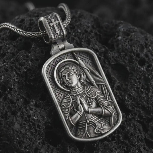 Oxidized Joan of Arc dog tag pendant necklace resting on black volcanic stone, featuring a detailed saint warrior portrait with halo, folded hands, and armor in a devotional sterling silver design.