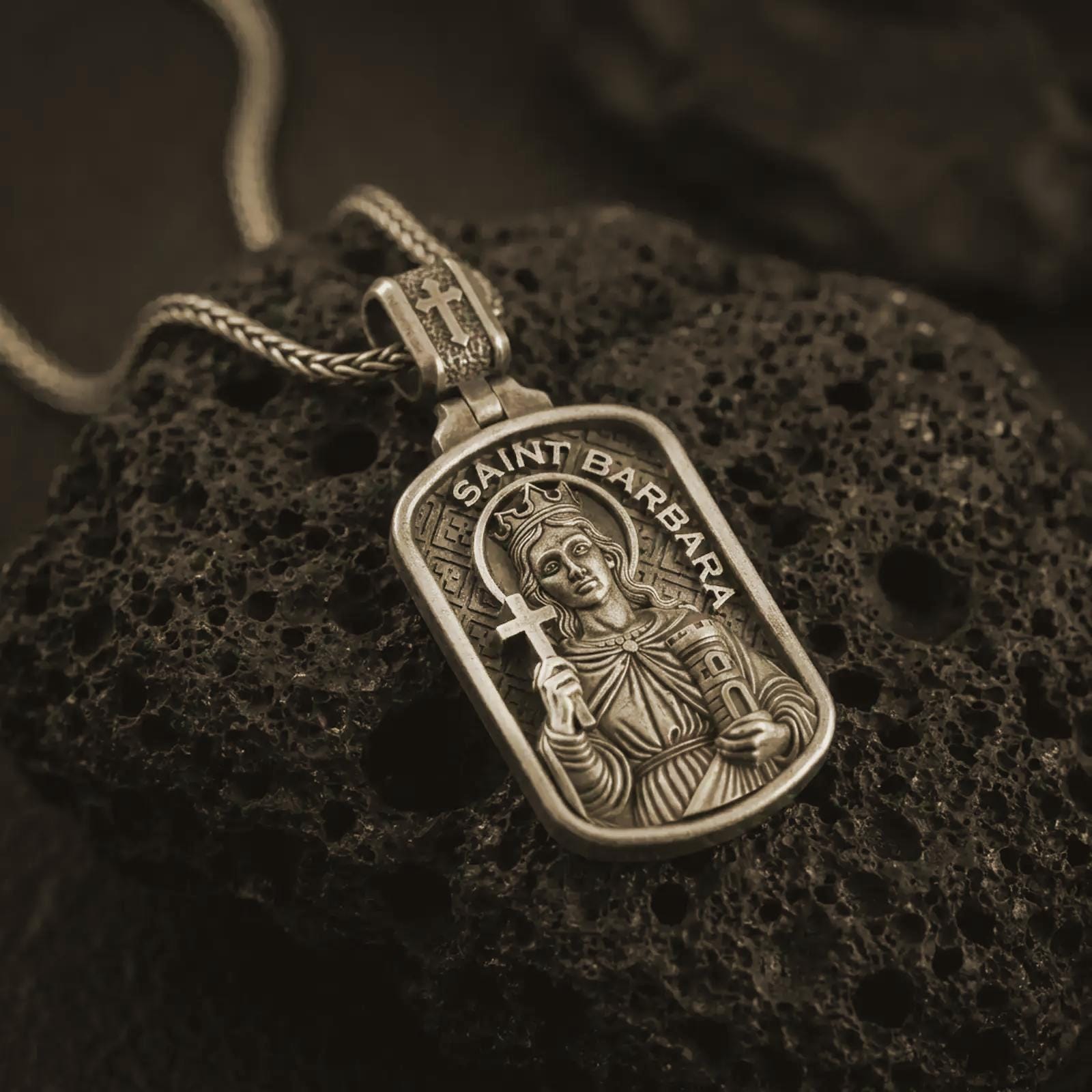 Bronze Saint Barbara pendant necklace on textured volcanic rock, close-up studio shot highlighting the saint relief, cross, tower detail, and dark antique silver contrast.