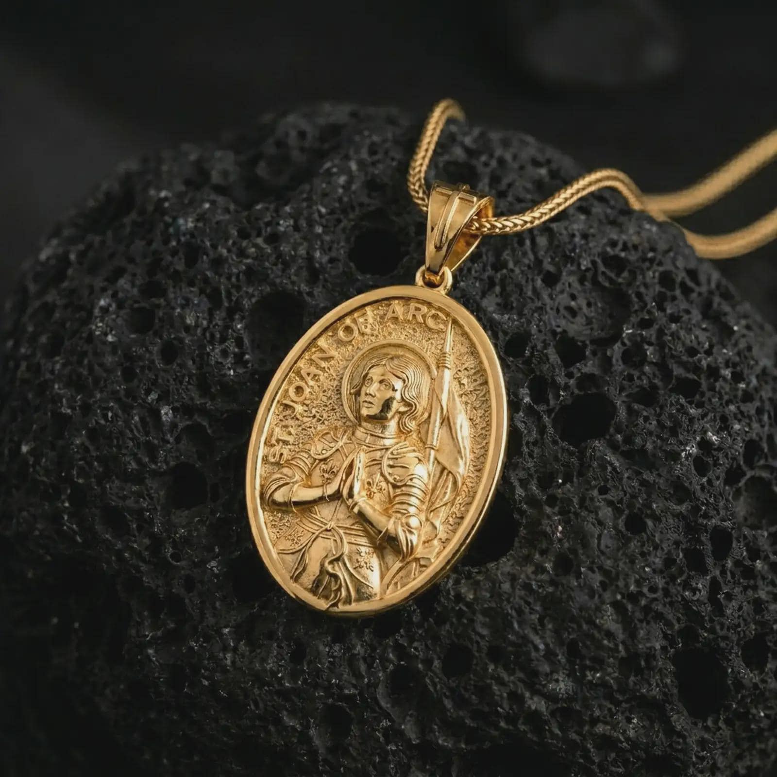 Gold plated Saint Joan of Arc pendant necklace on textured volcanic rock, premium close-up studio composition emphasizing the saint armor, prayer pose, and banner detail.