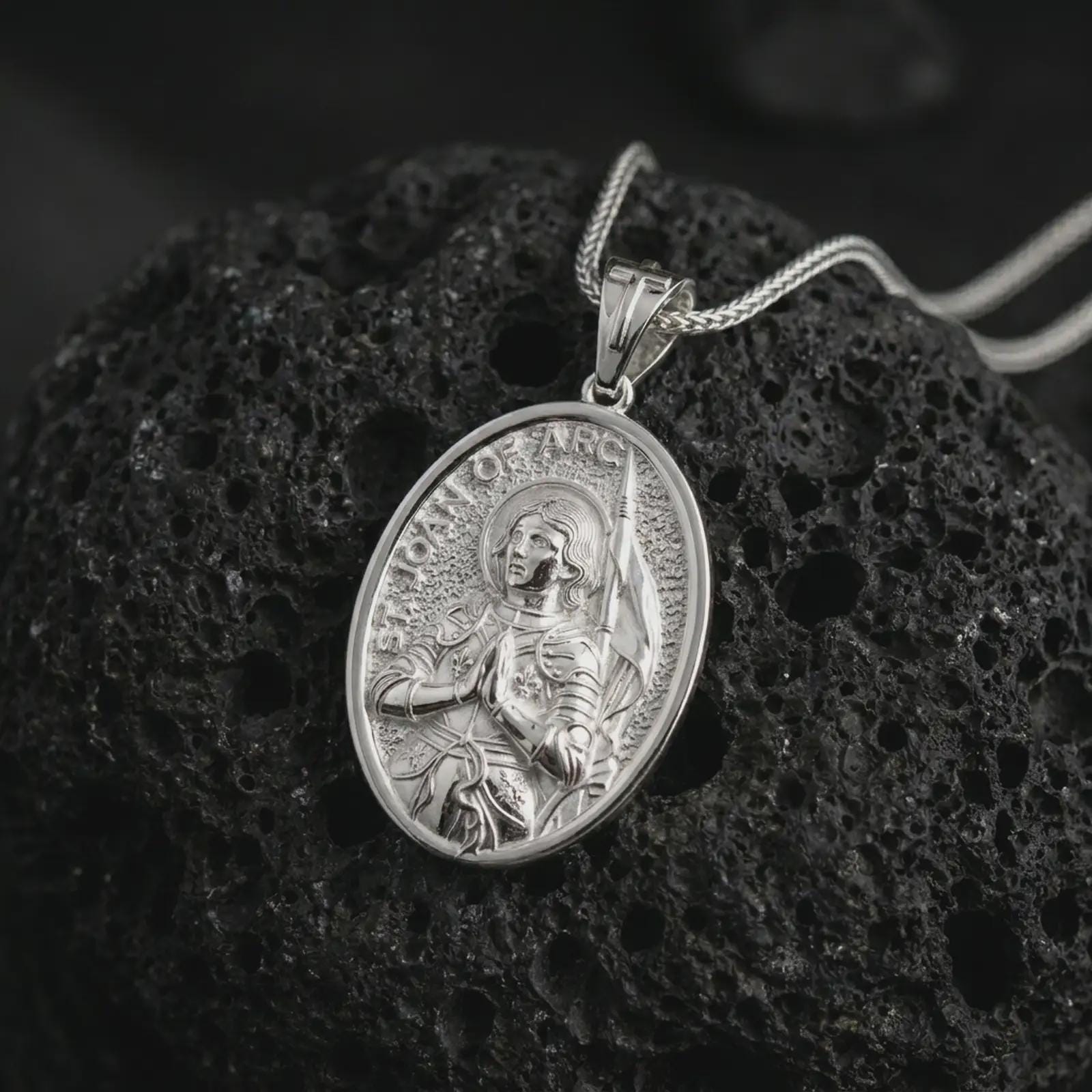 Polished silver Saint Joan of Arc pendant necklace on textured volcanic rock, centered macro jewelry photo with detailed Catholic medal engraving and bright silver highlights.