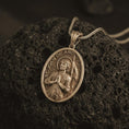 Load image into Gallery viewer, Bronze Saint Joan of Arc pendant necklace on textured volcanic rock, close-up studio shot highlighting the armored saint relief, halo, banner, and antique silver contrast.
