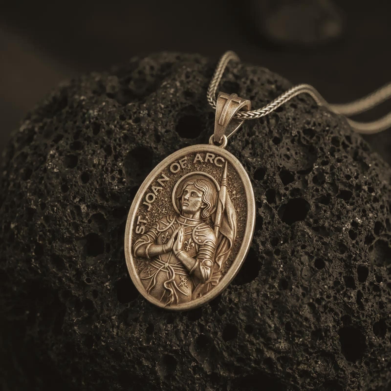 Bronze Saint Joan of Arc pendant necklace on textured volcanic rock, close-up studio shot highlighting the armored saint relief, halo, banner, and antique silver contrast.