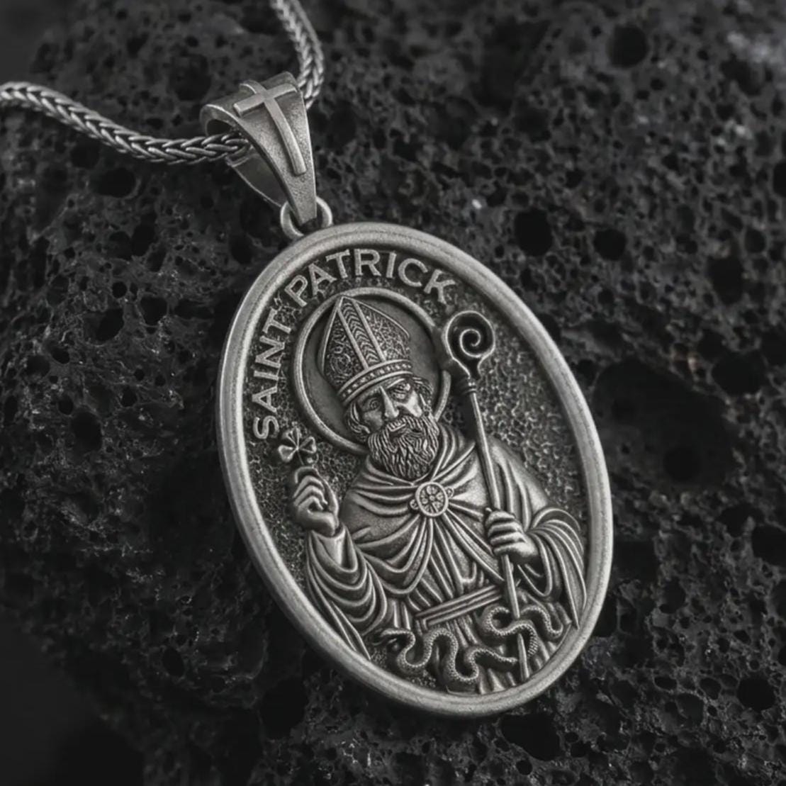 Oxidized Saint Patrick pendant necklace on textured volcanic rock, close-up studio shot highlighting the bishop relief, shamrock, staff, and antique silver contrast.