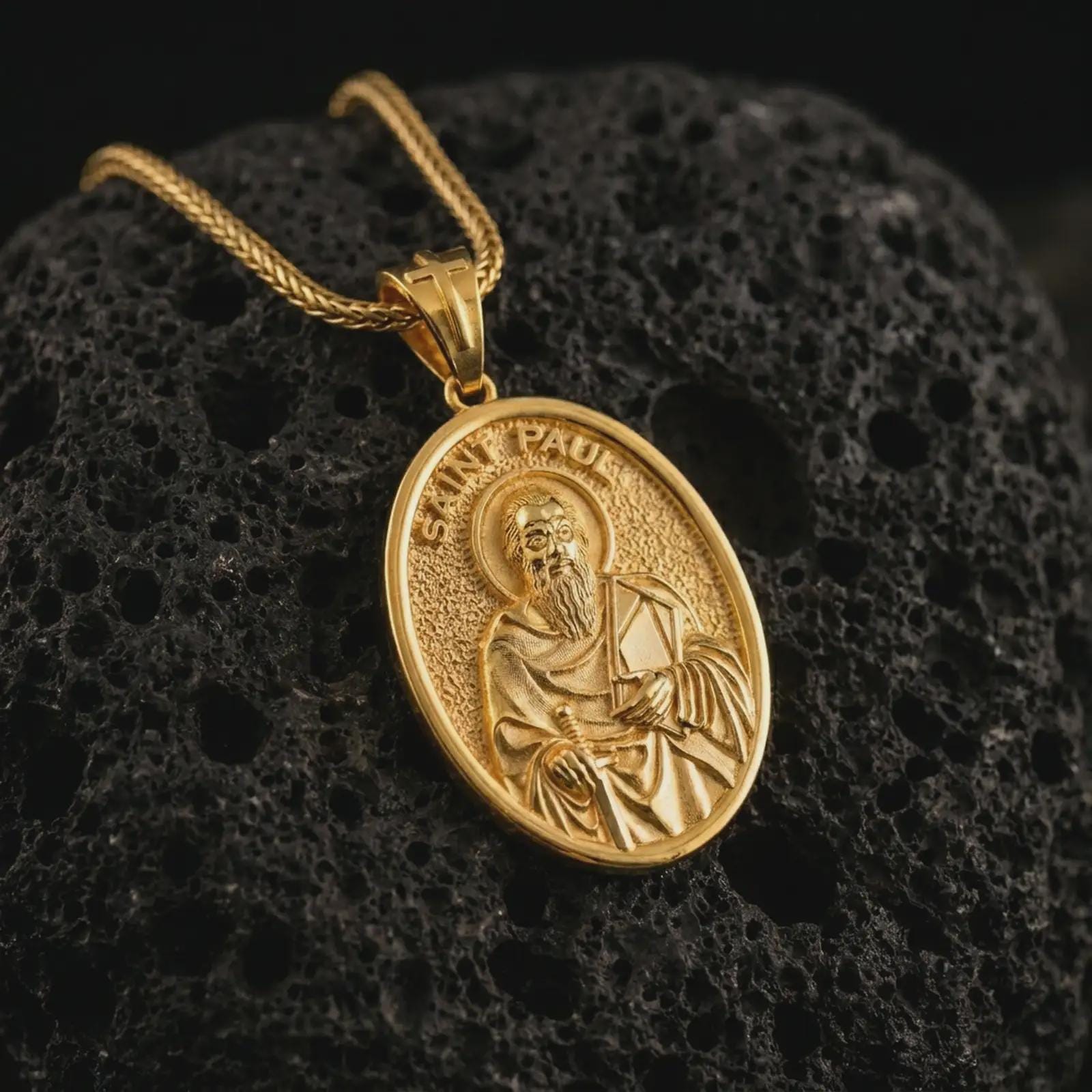 Gold plated Saint Paul pendant necklace on textured volcanic rock, premium close-up studio composition emphasizing the apostle figure, sword, and book symbolism.