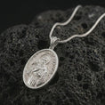 Load image into Gallery viewer, Polished silver Saint Francis pendant necklace on textured volcanic rock, centered macro jewelry photo with detailed religious engraving and bright silver highlights.
