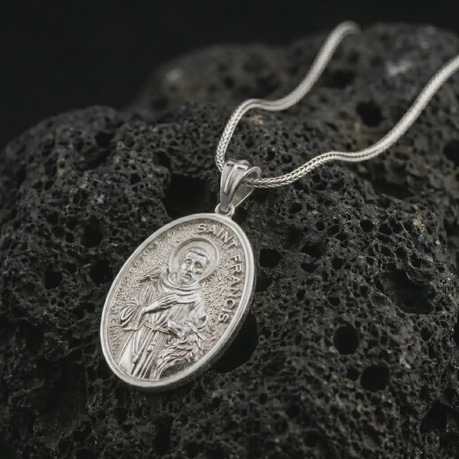 Polished silver Saint Francis pendant necklace on textured volcanic rock, centered macro jewelry photo with detailed religious engraving and bright silver highlights.