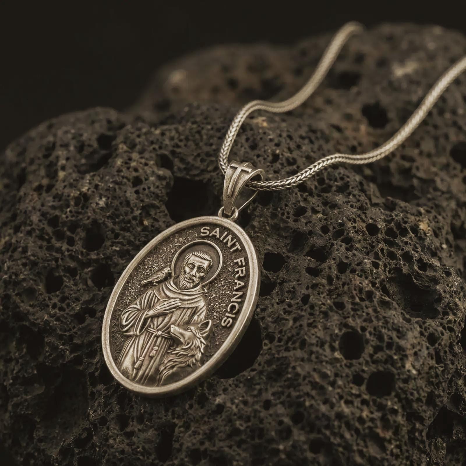 Bronze Saint Francis pendant necklace on textured volcanic rock, close-up studio shot highlighting the saint relief, bird, wolf detail, and antique silver contrast.