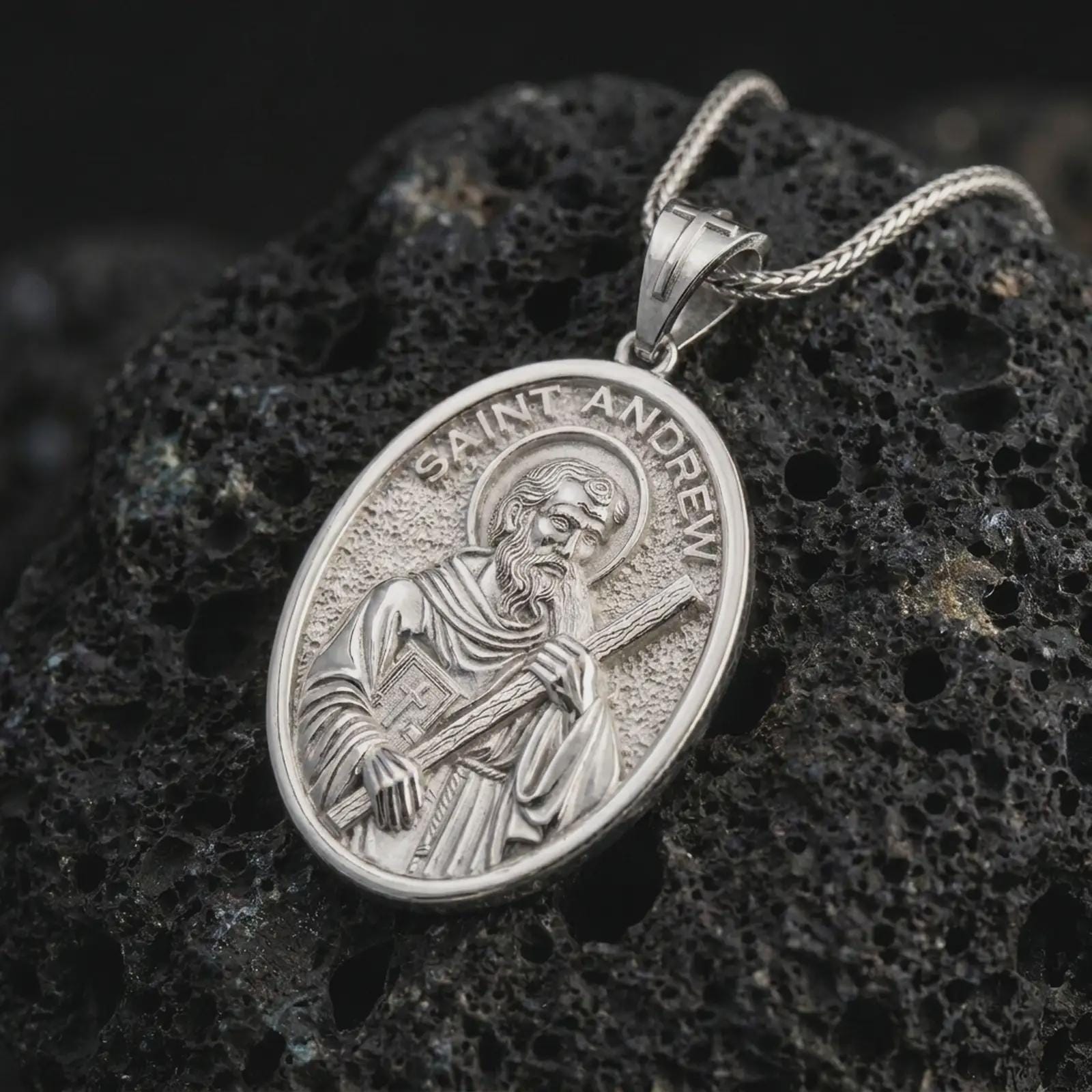 Polished silver Saint Andrew pendant necklace on textured volcanic rock, centered macro jewelry photo with detailed religious medal engraving and bright silver highlights.