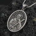 Load image into Gallery viewer, Oxidized Saint Andrew pendant necklace on textured volcanic rock, close-up studio shot highlighting the apostle relief, cross, book detail, and antique silver contrast.
