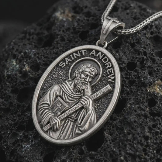 Oxidized Saint Andrew pendant necklace on textured volcanic rock, close-up studio shot highlighting the apostle relief, cross, book detail, and antique silver contrast.