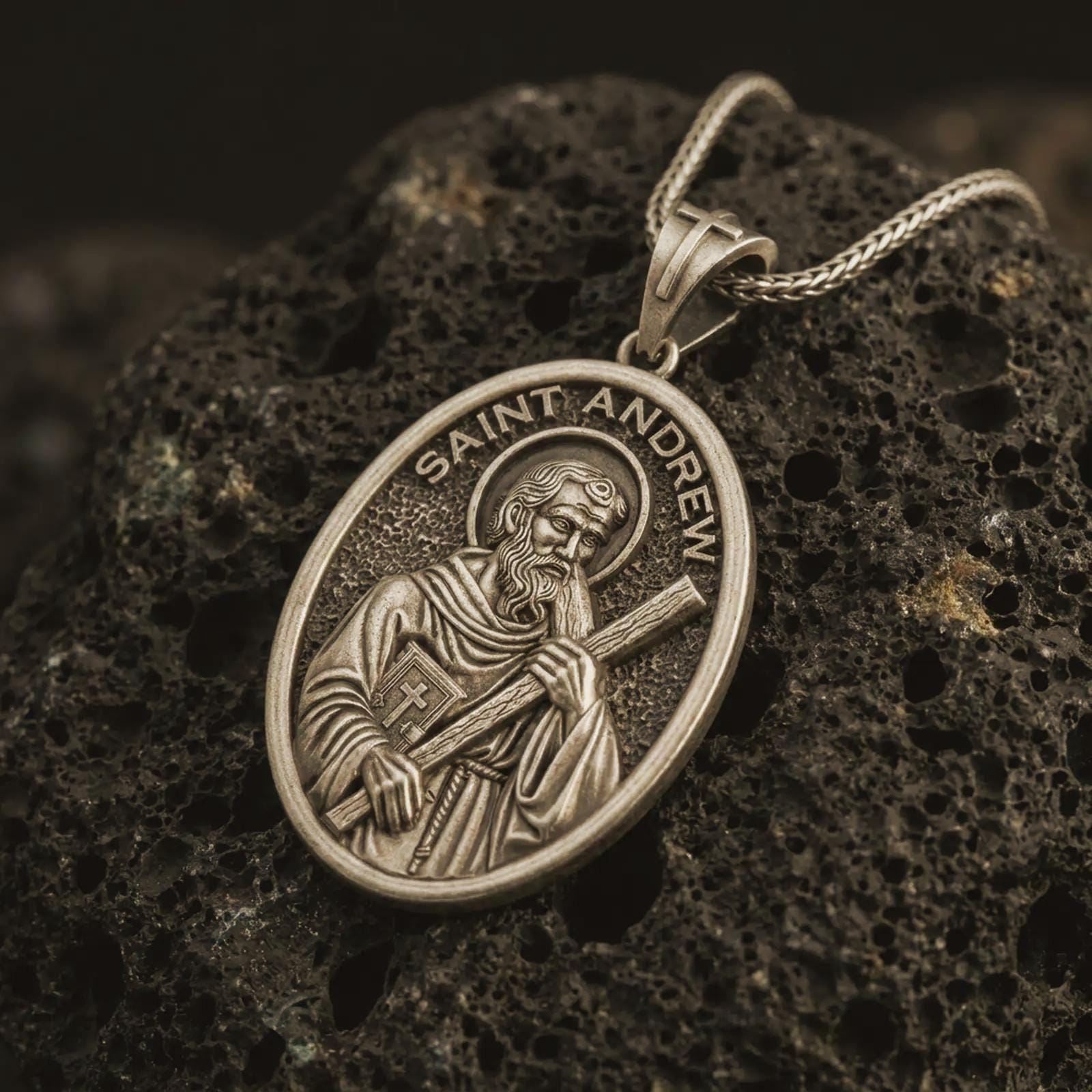 Bronze Saint Andrew pendant necklace on textured volcanic rock, close-up studio shot highlighting the apostle relief, cross, book detail, and antique silver contrast.