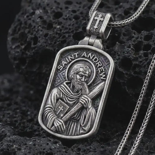 Oxidized Saint Andrew dog tag pendant necklace on textured volcanic rock, close-up studio shot highlighting the apostle relief, cross, book detail, and antique silver contrast.