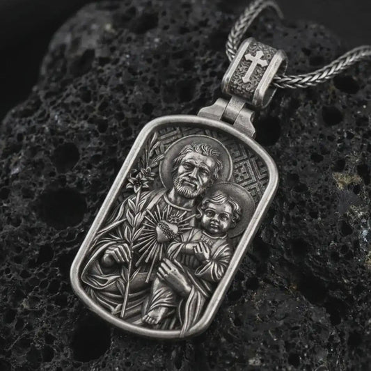 Oxidized Saint Joseph and Jesus pendant necklace on textured volcanic rock, close-up studio shot highlighting the father and child relief, lily, sacred heart, and antique silver contrast.
