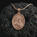 Load image into Gallery viewer, Rose gold plated Virgin Mary and baby Jesus pendant necklace on textured volcanic rock, detailed macro product shot with soft warm reflections and devotional mother-and-child symbolism.
